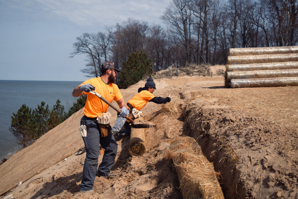 Two employees secure the erosion control blanket at the top of the dune, using trenching and staking in place.